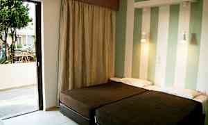 Kaksi yhden hengen vuodetta huoneessa, jossa on raidalliset seinät ja avoin parvekkeen ovi, joka johtaa ulkoterassille. (Tekoälyn luoma teksti)