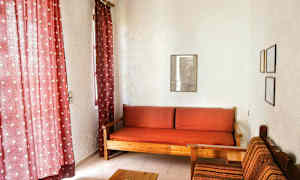 Olohuoneessa on punaiset verhot, puusohva punaisilla ruudullisilla tyynyillä ja kangaspäällysteinen tuoli vieressä. (Tekoälyn luoma teksti)