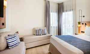 Moderni hotellihuone, jossa on sänky, sohva ja ikkunat vaaleilla verhoilla. (Tekoälyn luoma teksti)