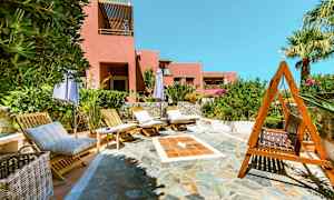 Aurinkotuoleja ja aurinkovarjoja kivetyllä terassialueella, jossa taustalla vaaleanpunainen rakennus. (Tekoälyn luoma teksti)