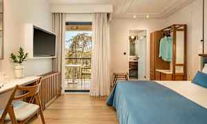 Hotellihuone, jossa on parivuode, työpöytä ja televisio. Lasiovet johtavat parvekkeelle. Kylpyhuone näkyy avoimen oven läpi. (Tekoälyn luoma teksti)