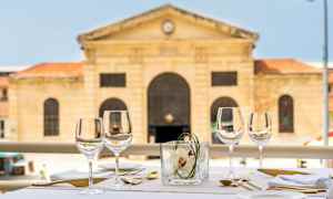 Ulkona katettu pöytä viinilaseilla, ruokailuvälineillä ja lautasliinoilla. Taustalla näkyy historiallinen kivirakennus. (Tekoälyn luoma teksti)