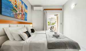 Kahden hengen huone, jossa on valkoiset seinät, parivuode harmaalla päiväpeitteellä ja seinällä purjeveneaiheinen maalaus. (Tekoälyn luoma teksti)