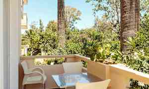 Parveke, jossa on pöytä ja tuoleja, ympäröitynä vihreillä kasveilla ja palmuilla. (Tekoälyn luoma teksti)