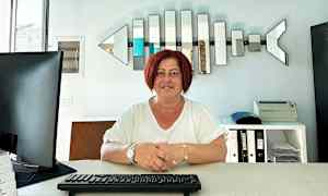 Henkilö istuu työpöydän ääressä, jossa on näppäimistö, tietokonenäyttö ja hyllyjä taustalla. (Tekoälyn luoma teksti)