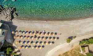 Ilmakuva rannasta, jossa on aseteltuja aurinkotuoleja ja varjoja turkoosin veden reunalla. (Tekoälyn luoma teksti)