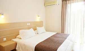 Hotellihuone, jossa on suuri sänky, valkoiset vuodevaatteet ja ruskea peitto. Seinälle on asennettu ilmastointilaite ikkunan vieressä, jossa on vaaleat verhot. (Tekoälyn luoma teksti)