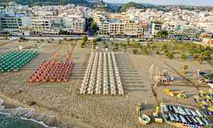 Ranta, jossa on riveissä aurinkovarjoja ja aurinkotuoleja, kaupungin ja monien rakennusten edessä. (Tekoälyn luoma teksti)