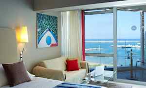 Hotellihuone, jossa oleskelutila, seinällä valomaalaus ja lasi-ikkunat, joista näkyy meri ja pieni veneilysatama. (Tekoälyn luoma teksti)