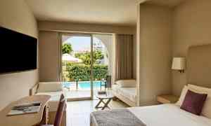 Hotellihuone, jossa on parivuode, kirjoituspaikka ja lisävuode. Ulkona näkyy terassi ja uima-allas. (Tekoälyn luoma teksti)