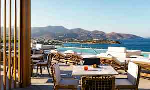 Terassi, jolta on näkymä merelle ja vuorille taustalla; mukana aurinkotuoleja ja pöytiä. (Tekoälyn luoma teksti)