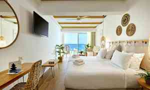 Hotellihuone, jossa on valkoiset seinät, pedattu sänky, istuimia ja parveke merinäköalalla. (Tekoälyn luoma teksti)