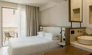 Hotellihuoneen sisustus, jossa on parivuode, ikkuna verhoineen, parveke tuolilla ja pesuallas. (Tekoälyn luoma teksti)