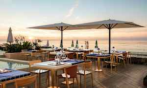 Meren rannalla sijaitseva ulkoilmaravintola auringonlaskun aikaan, pöydillä ja tuoleilla sekä suurilla aurinkovarjoilla. (Tekoälyn luoma teksti)