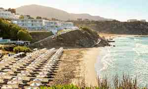 Ranta, jossa aurinkotuoleja ja varjoja, rakennuksia rinteessä. (Tekoälyn luoma teksti)