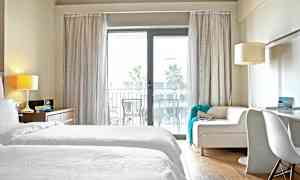 Hotellihuone, jossa valkoiset vuodevaatteet ja sohva lasioven edessä parvekkeelle. (Tekoälyn luoma teksti)