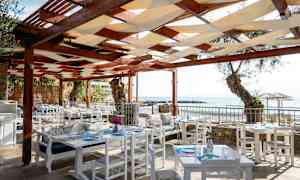 Ulkoravintola valkoisilla pöydillä ja tuoleilla puisen katon alla. Merimaisema taustalla. (Tekoälyn luoma teksti)