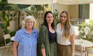 Kolme henkilöä seisoo ulkona pöydän ja tuolien vieressä aurinkovarjon alla. (Tekoälyn luoma teksti)