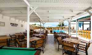 Ulkoravintola, jossa on useita puupöytiä ja tuoleja valkoisen katon alla lähellä ulkouima-allasta. (Tekoälyn luoma teksti)