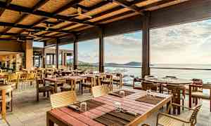 Avoin ja valoisa ravintola, jossa on puupöydät ja tuolit, merinäkymällä horisonttiin. (Tekoälyn luoma teksti)