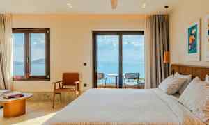 Hotellihuone, jossa petaettu sänky, tuoleja ja pöytä ikkunan ääressä, merinäköala. (Tekoälyn luoma teksti)