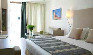 Moderni hotellihuone, jossa suuri sänky, työpöytä ja mukava istuinvaunu vaaleissa väreissä. (Tekoälyn luoma teksti)