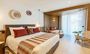 Hotellihuone, jossa on parisänky, koristeltu punaisilla ja beigeillä tekstiileillä, ja oleskelutila ikkunan ääressä luonnonvalolla. (Tekoälyn luoma teksti)