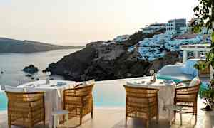 Ulkoterassi, jossa on pöytiä ja tuoleja altaan äärellä, maisemana rannikko ja valkoisia rakennuksia kukkulalla. (Tekoälyn luoma teksti)