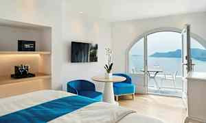 Hotellihuone, jossa on vaaleat valkoiset seinät, kalustettu sängyllä, pöydällä ja tuoleilla, sekä parveke, jolta on näkymä merelle ja saarelle. (Tekoälyn luoma teksti)