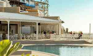 Ulkoterassi, jossa valkoiset huonekalut altaan vieressä ja muutama henkilö istumassa pöytien ääressä. Taustalla rakennuksia ja vaalea taivas. (Tekoälyn luoma teksti)