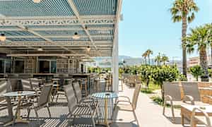 Ulkoiluterassi, jossa on pöytiä ja tuoleja kattorakenteen alla, ympärillä vehreyttä ja palmuja. (Tekoälyn luoma teksti)