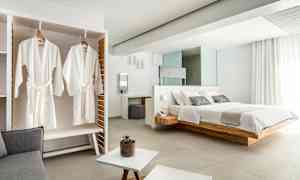 Kuva tyylikkäästä hotellihuoneesta, jossa on suuri sänky, vaatekaapissa roikkuvat valkoiset kylpytakit ja moderneja vaaleita huonekaluja. (Tekoälyn luoma teksti)