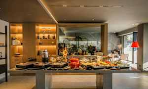 Buffet-alue, jossa erilaisia ruokia ja juomia pöydällä. Tausta, jossa hyllyjä ja istumapaikkoja. (Tekoälyn luoma teksti)