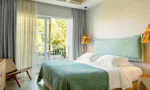 Hotellihuone, jossa on parivuode, siniset vuodevaatteet ja suuret ikkunat, jotka avautuvat vehreälle parvekkeelle. (Tekoälyn luoma teksti)