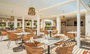 Ulkoravintola, jossa on istuinpaikkoja ja pöytiä, valkoisilla pergolapalkkeilla ja koristeltu rottinkilampuilla. Baari näkyy taustalla. (Tekoälyn luoma teksti)