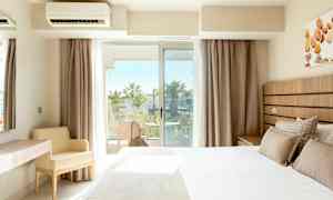 Hotellihuone, jossa on sänky, kirjoituspöytä ja tuoli, sekä parveke, josta näkyy palmuja. (Tekoälyn luoma teksti)