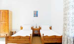 Kaksi erillistä sänkyä puisilla kehyksillä valoisassa huoneessa, jonka seinät ovat valkoiset. Pieni yöpöytä ja lamppu niiden välissä. (Tekoälyn luoma teksti)