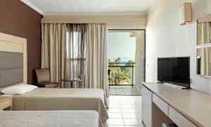 Hotellihuone, jossa on kaksi sänkyä, TV, pöytä ja tuoleja sekä parveke näkymällä vehreään maisemaan ja merelle. (Tekoälyn luoma teksti)