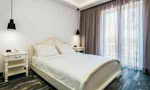 Makuuhuone, jossa on valkoinen sänky ja valkoiset yöpöydät, suuren ikkunan vieressä vaaleilla verhoilla. (Tekoälyn luoma teksti)