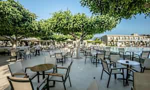 Ulkotila, jossa tuoleja ja pöytiä puiden alla lähellä allasta. (Tekoälyn luoma teksti)