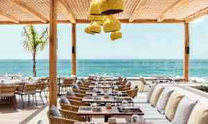 Ravintola merinäköalalla, jossa puisia ja rottinkisia pöytiä ja tuoleja sekä tyynyillä varustettuja penkkejä suojaisan katon alla. (Tekoälyn luoma teksti)