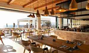 Ravintola, jossa puisia pöytiä, tuoleja ja koristeellisia valaisimia, ja näkymä merelle. (Tekoälyn luoma teksti)