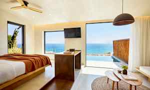 Huone, josta on näkymä merelle suurten ikkunoiden läpi, vasemmalla on sänky ja etualalla pöytä, jossa on esineitä. (Tekoälyn luoma teksti)