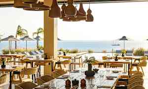 Ravintola, jossa on pöytiä ja tuoleja, näkymä ulkouima-altaalle, merelle ja aurinkovarjoille. (Tekoälyn luoma teksti)