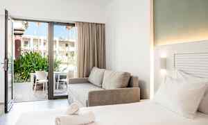 Moderni hotellihuone, jossa on parivuode, harmaa sohva ja avoin lasiovi, joka johtaa terassille, jolla on tuoleja ja vihreää kasvillisuutta. (Tekoälyn luoma teksti)