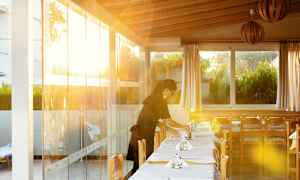 Ravintola, jossa pöydät ovat katettuina, ja auringonvalo paistaa lasiseinien läpi, ruukkukukkia näkyy ikkunan ulkopuolella. (Tekoälyn luoma teksti)