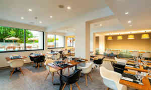 Moderni ravintola, jossa on puukalusteet, ikkunat näkymällä ulkoterassille ja useita pöytäkattauksia. (Tekoälyn luoma teksti)