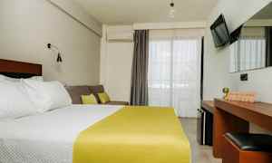 Hotellihuone, jossa on parivuode, valkoiset vuodevaatteet ja keltainen päiväpeite, sohva tyynyineen, työpöytä peileineen ja seinälle kiinnitetty TV. Parvekkeen ovet vaaleilla verhoilla. (Tekoälyn luoma teksti)