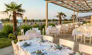 Ulkoravintola, jossa on valkoisia pöytiä ja tuoleja, palmuja ja merinäköala. (Tekoälyn luoma teksti)