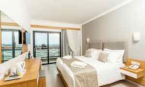 Hotellihuone, jossa iso sänky, vaaleat kalusteet ja parveke näkymällä. (Tekoälyn luoma teksti)